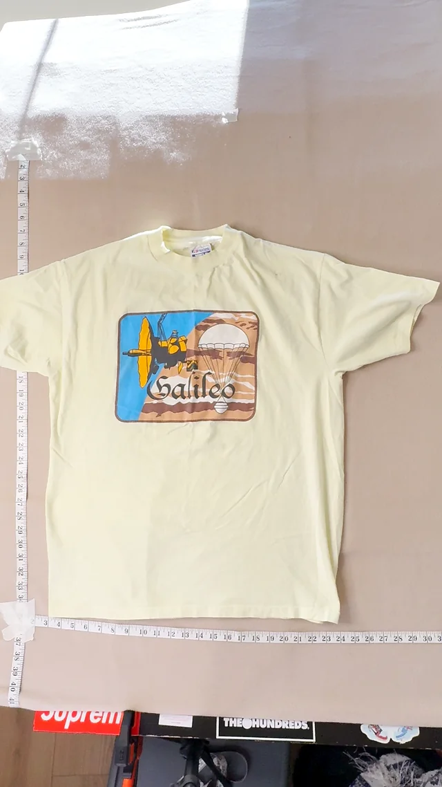 Original front frame: a pale yellow vintage Galileo Spacecraft t-shirt laid flat on a tan backdrop with tape measures and clutter around it.