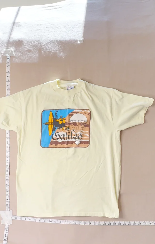 Original front photo straight from the video: the Galileo tee on a tan backdrop with measuring tapes taped along the edges and glare on the cloth.